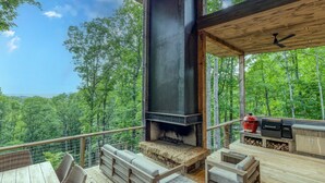 An outdoor deck with a fireplace, seating, and a grill, overlooking a forested landscape. - Greenridge - Hot tub & mountain views (Blue Ridge Mountains)