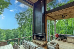 An outdoor deck with a fireplace, seating, and a grill, overlooking a forested landscape.