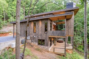 A two-story wooden cabin with a deck, surrounded by forest.