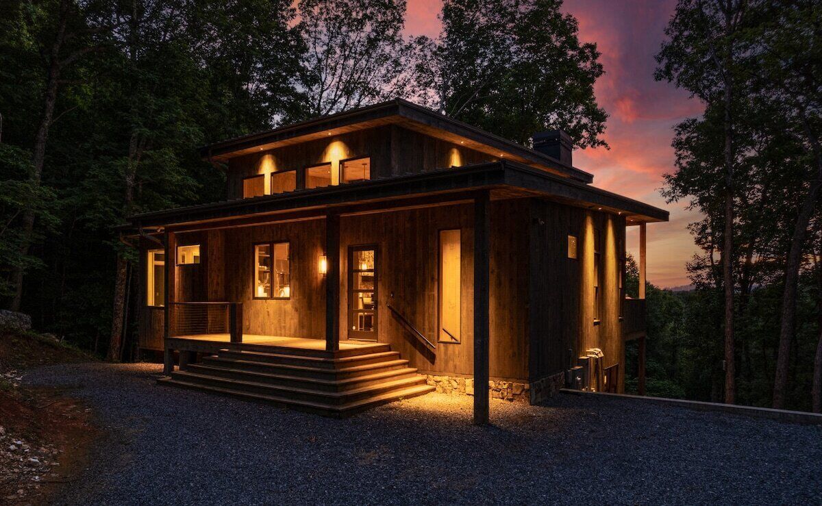 A wooden cabin with a porch, steps leading to the entrance, and a view of the surrounding forest.