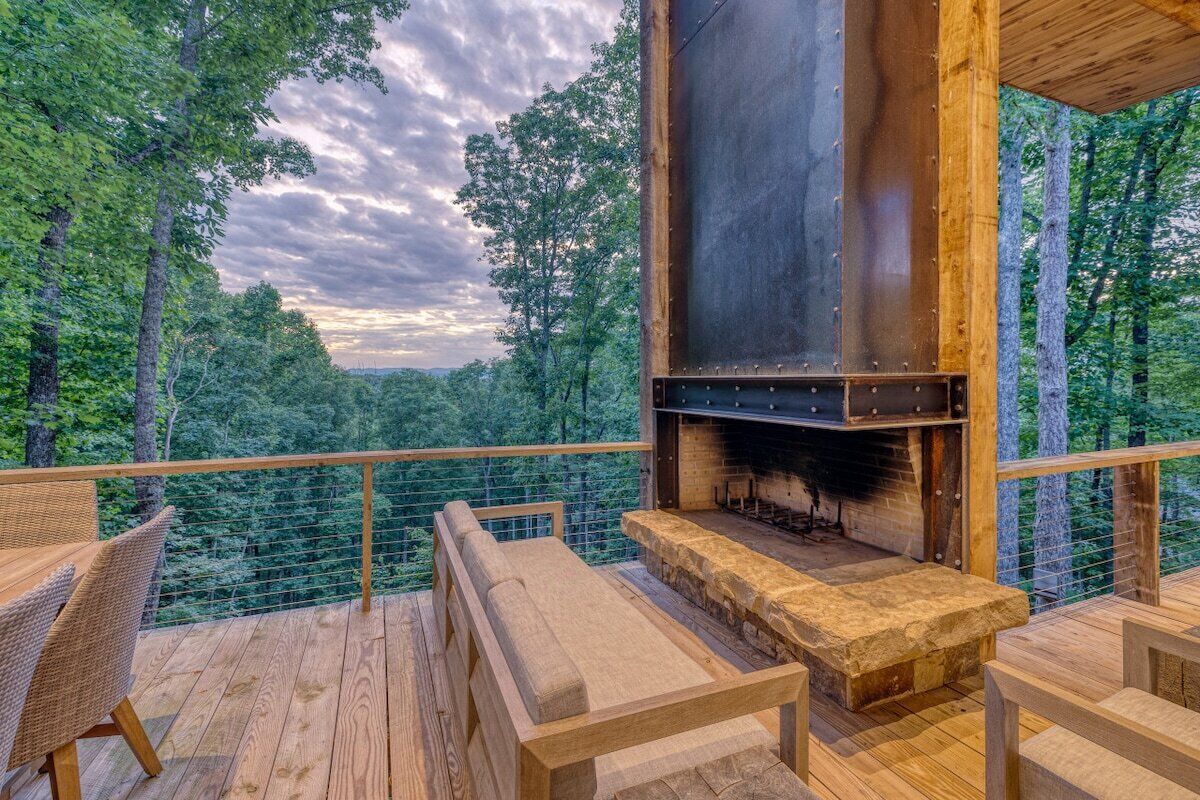An outdoor deck with a stone fireplace, wooden furniture, and a view of a forested landscape.