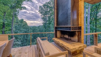 An outdoor deck with a stone fireplace, wooden furniture, and a view of a forested landscape.