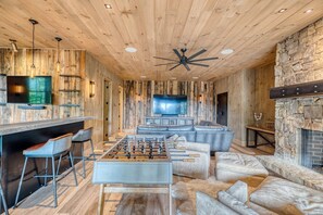 A room with a wooden ceiling, a ceiling fan, a foosball table, a television, and a stone fireplace.