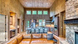A spacious living area with a leather sofa, a stone fireplace, and a kitchen in the background. - Greenridge - Hot tub & mountain views (Blue Ridge Mountains)
