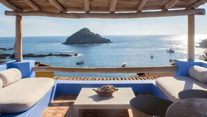 Une terrasse offrant une vue sur la mer, agrémentée d’une pergola en bois, de sièges blancs dotés de coussins et d’un sol carrelé.
