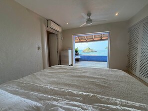 A bedroom with a bed, an air conditioning unit, a ceiling fan, and a view of the ocean through an open door.