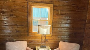 A cozy room with wooden walls, two patterned armchairs, a small table with a lamp, and a plaid rug. - #29 - Flagpole View Cabins - Deluxe Cabin (Columbia Falls)