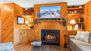 A cozy living room with a fireplace, wooden walls, a sofa, and a television mounted above the fireplace.