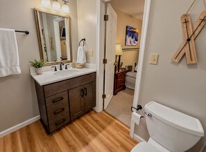 Un baño con piso de madera, inodoro blanco, tocador con espejo y un perchero para toallas.