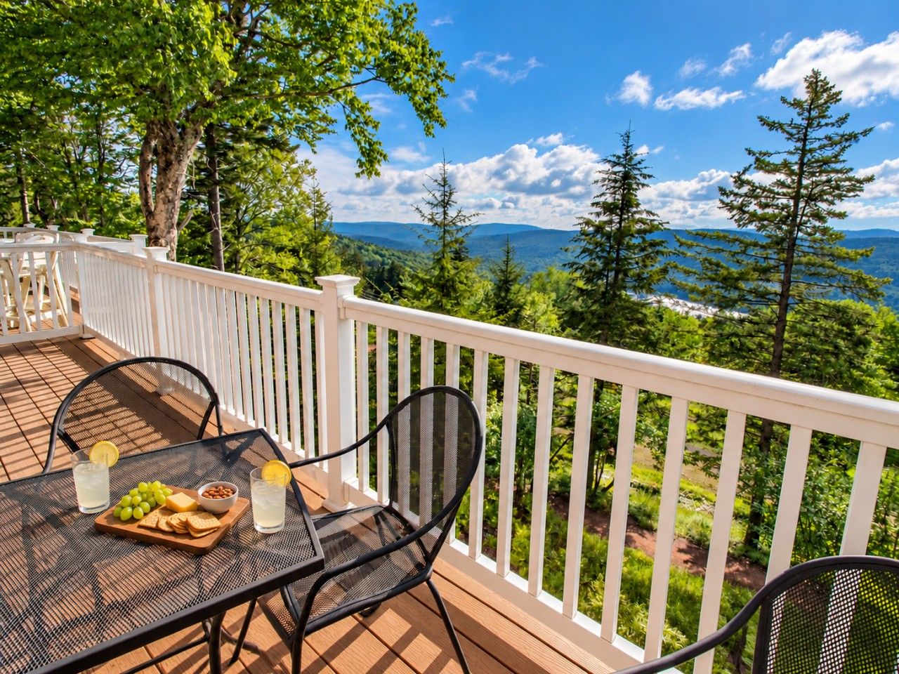 Une terrasse avec une table dressée pour deux personnes, sur laquelle on trouve une planche avec des raisins, des crackers et un bol de noix, ainsi que deux verres contenant une boisson claire.