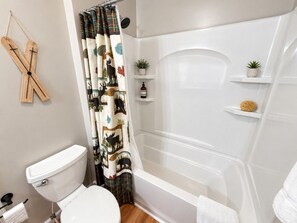 Un baño con inodoro, ducha y cortina decorada con motivos naturales. Hay estantes con artículos de aseo personal y una pequeña planta.