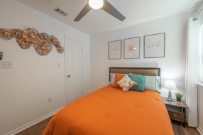A bedroom with an orange bedspread, a wooden headboard, and a ceiling fan. There are three framed pictures on the wall and a nightstand with a lamp and a plant.