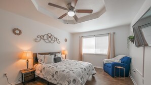 Une chambre à coucher avec un lit, deux lampes de chevet, un ventilateur de plafond, une fenêtre avec des rideaux, une chaise bleue et une petite table.