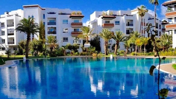 Una piscina rodeada de edificios modernos con balcones, palmeras y un cielo azul despejado.