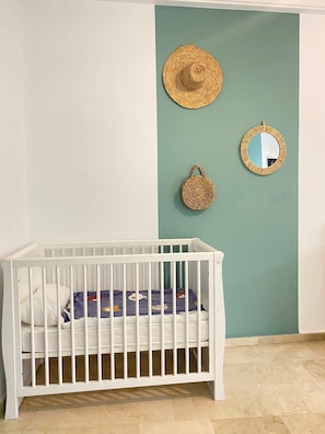 Un berceau blanc avec un matelas à motifs, placé contre un mur de couleur turquoise, accompagné d’un panier en osier et d’un miroir rond dont le cadre est également en osier.