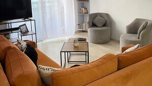 Una sala moderna con un sofá grande naranja, un sillón gris, una mesita de centro pequeña y un televisor de pantalla plana sobre un soporte.