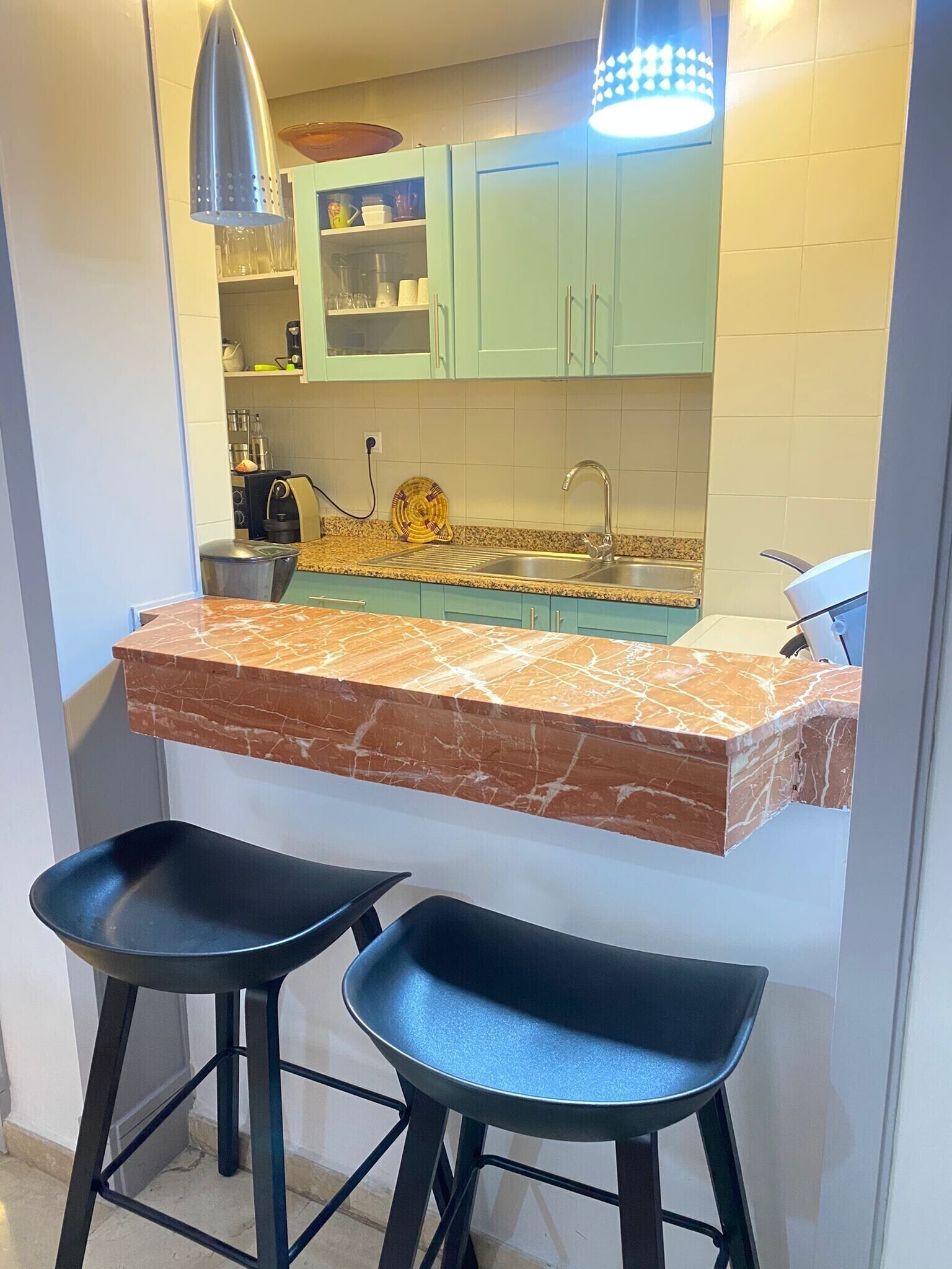 A compact kitchen with a marble countertop, two black bar stools, and light green cabinets.