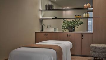 A modern hotel room with a large mirror, a bedside table, a plant, and a patterned ottoman.
