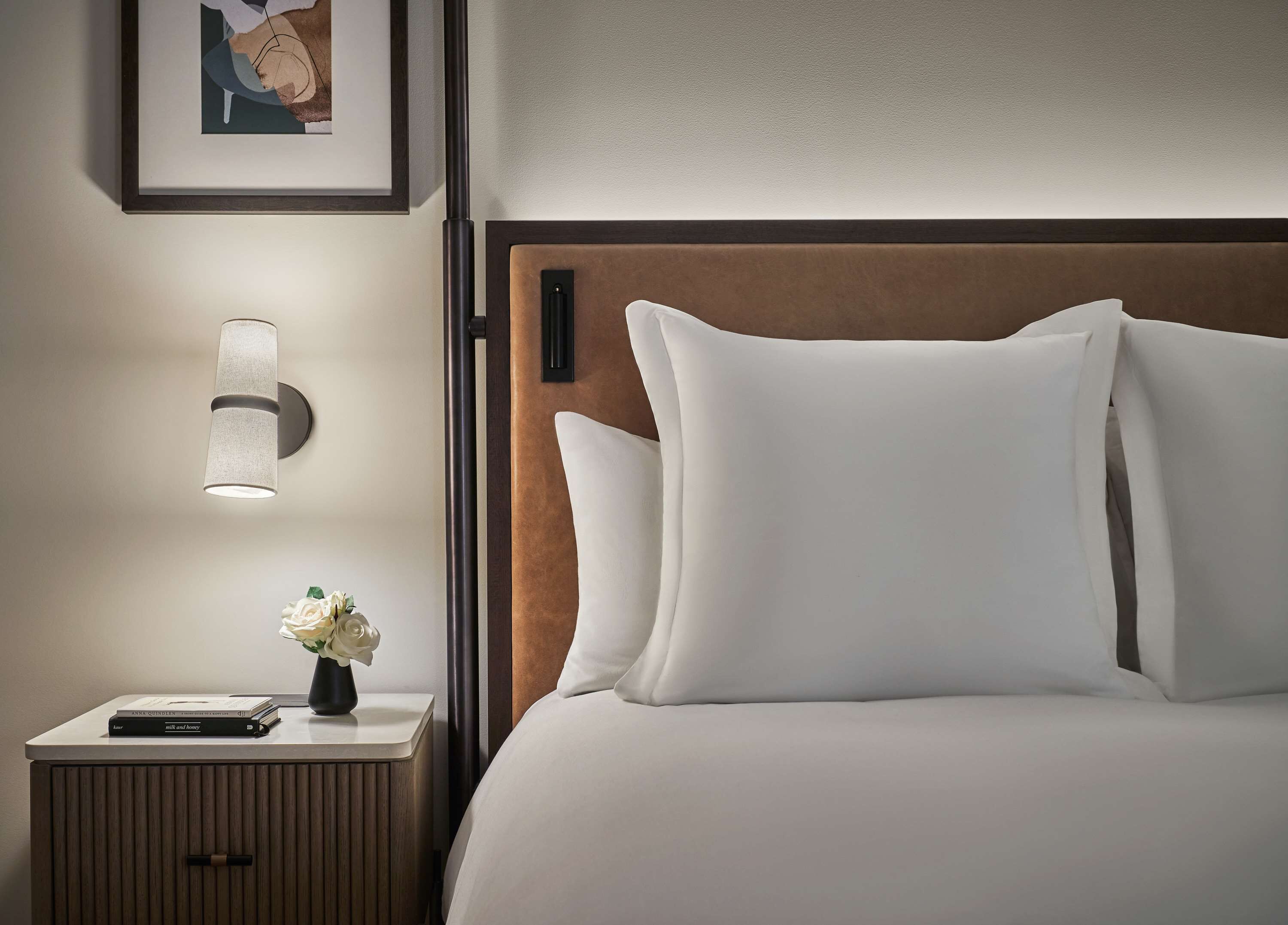 Een net opgemaakt bed met witte lakens, een bedlamp, een ingelijst schilderij aan de muur, en een klein tafeltje met een bloevas en boeken.
