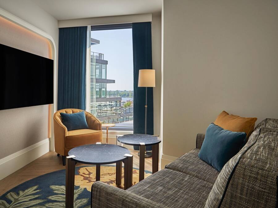 Ein modernes Hotelzimmer mit großem Fenster, einem an der Wand montierten Flachbildfernseher, einer Couch, zwei kleinen runden Tischen und einer Lampe.