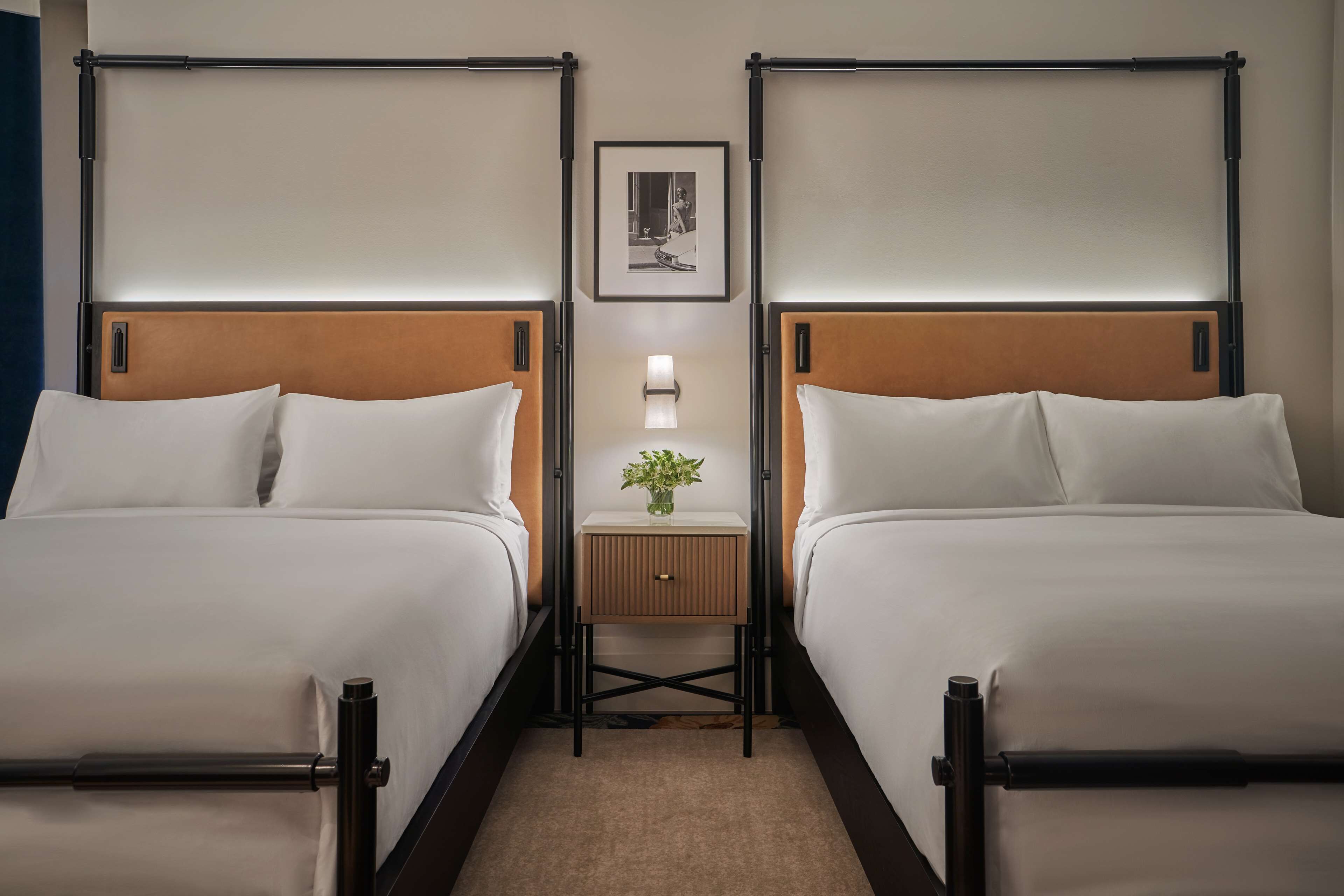 Habitación de hotel con dos camas, una mesita de noche con lámpara y un cuadro enmarcado en la pared.