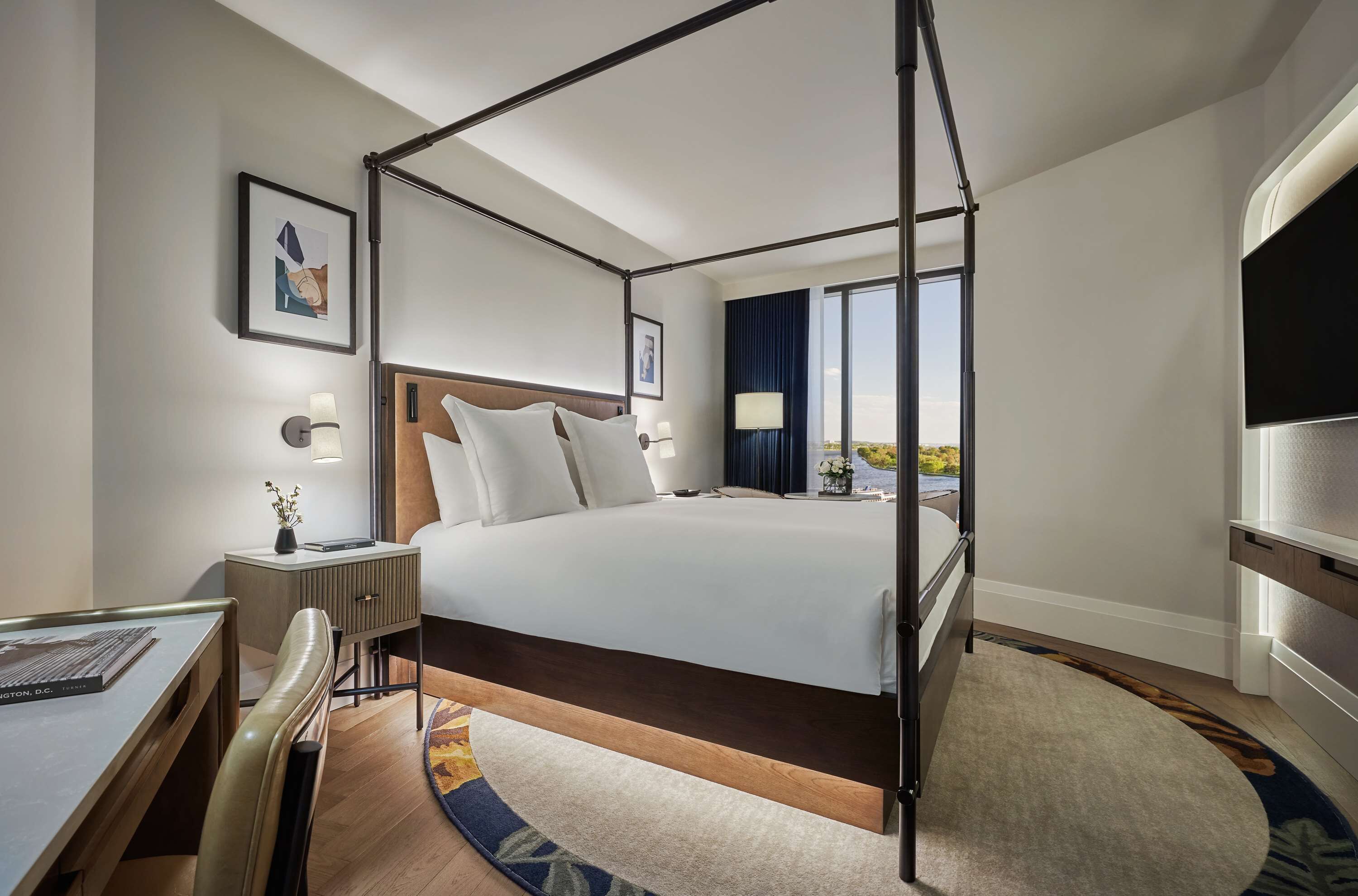 A modern hotel room with a four-poster bed, a desk with a chair, a flat-screen TV, and a large window offering a view of the outdoors.