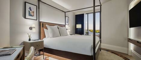 A modern hotel room with a four-poster bed, a desk with a chair, a flat-screen TV, and a large window offering a view of the outdoors.