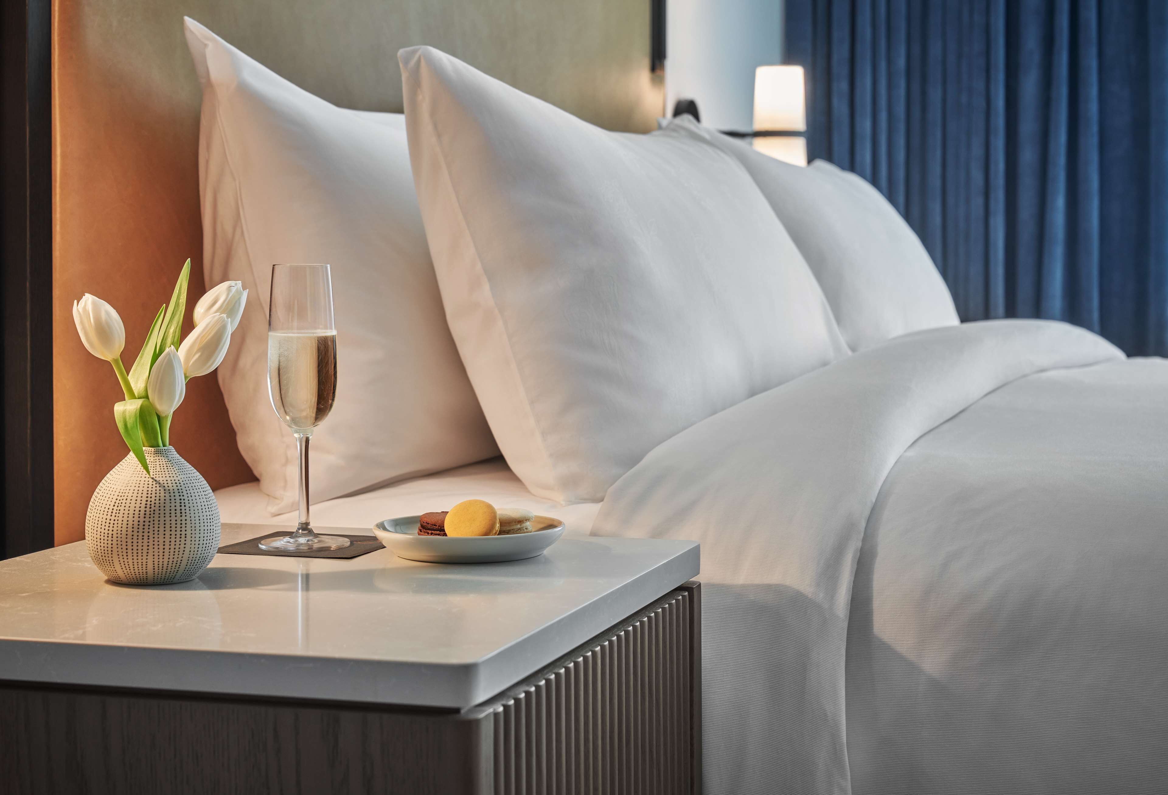 A neatly made bed with white linens, a small table with a vase of white tulips, a glass of champagne, and a plate of cookies.