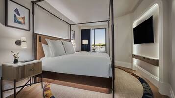 A modern hotel room with a large bed, a bedside table with a lamp, a flat-screen TV mounted on the wall, and a view of the outdoors.