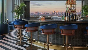 A modern bar with a city view, featuring hanging pendant lights, blue bar stools, and a central liquor display. - Pendry Washington DC - The Wharf (Washington)