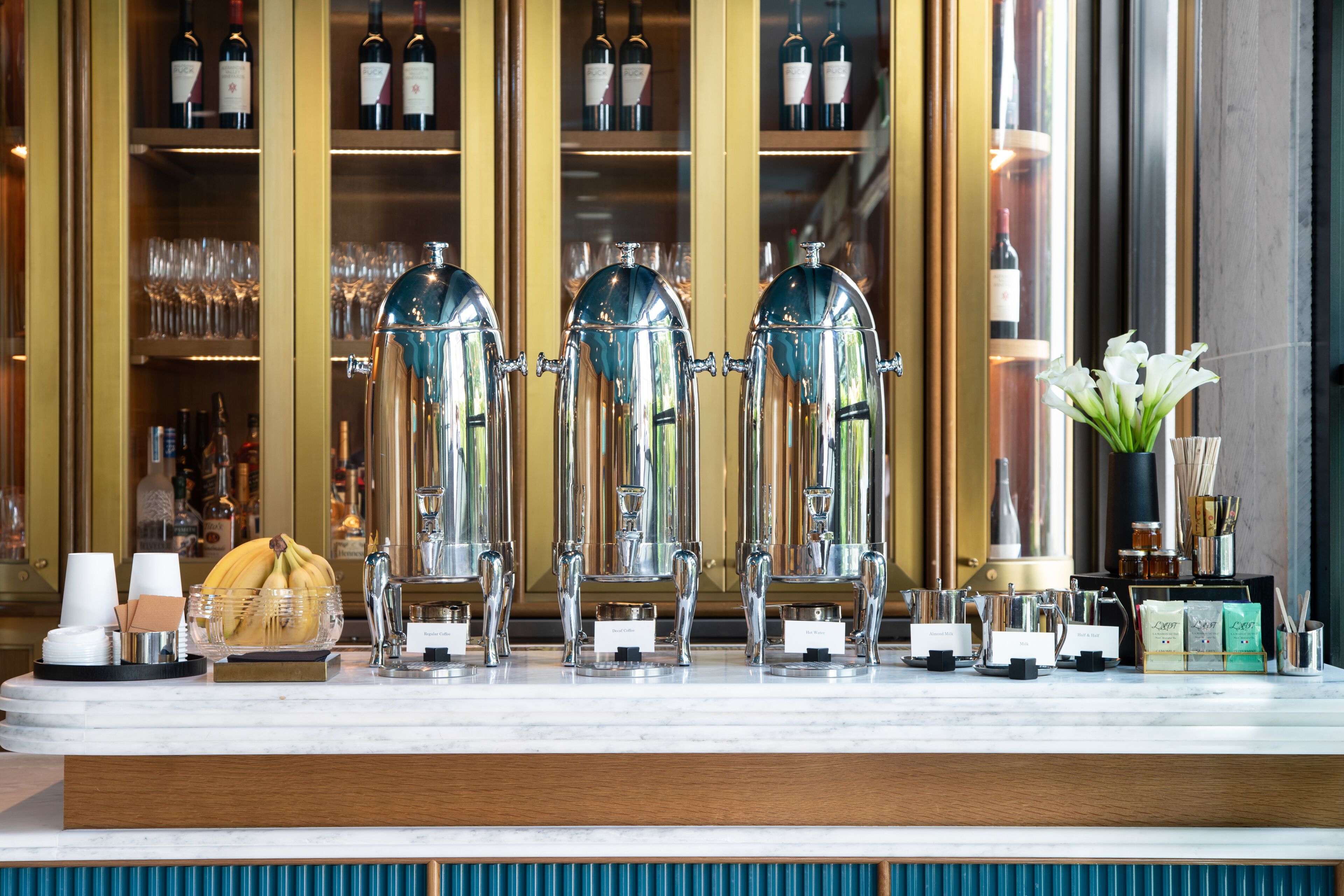 A bar area with three coffee machines, a vase with white flowers, and shelves with wine bottles and glasses.