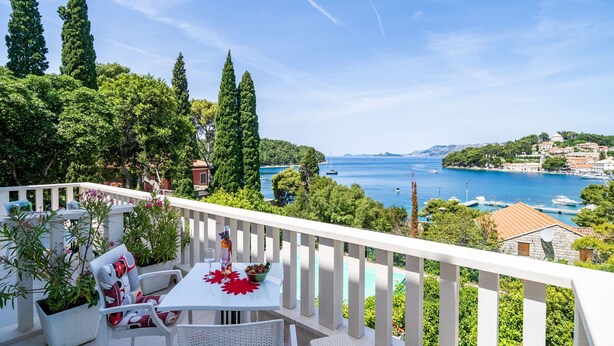 Ein Balkon mit weißen Korbmöbeln, einem Tisch mit roter Dekoration und Blick auf eine Küstenstadt mit Booten und viel Grün.