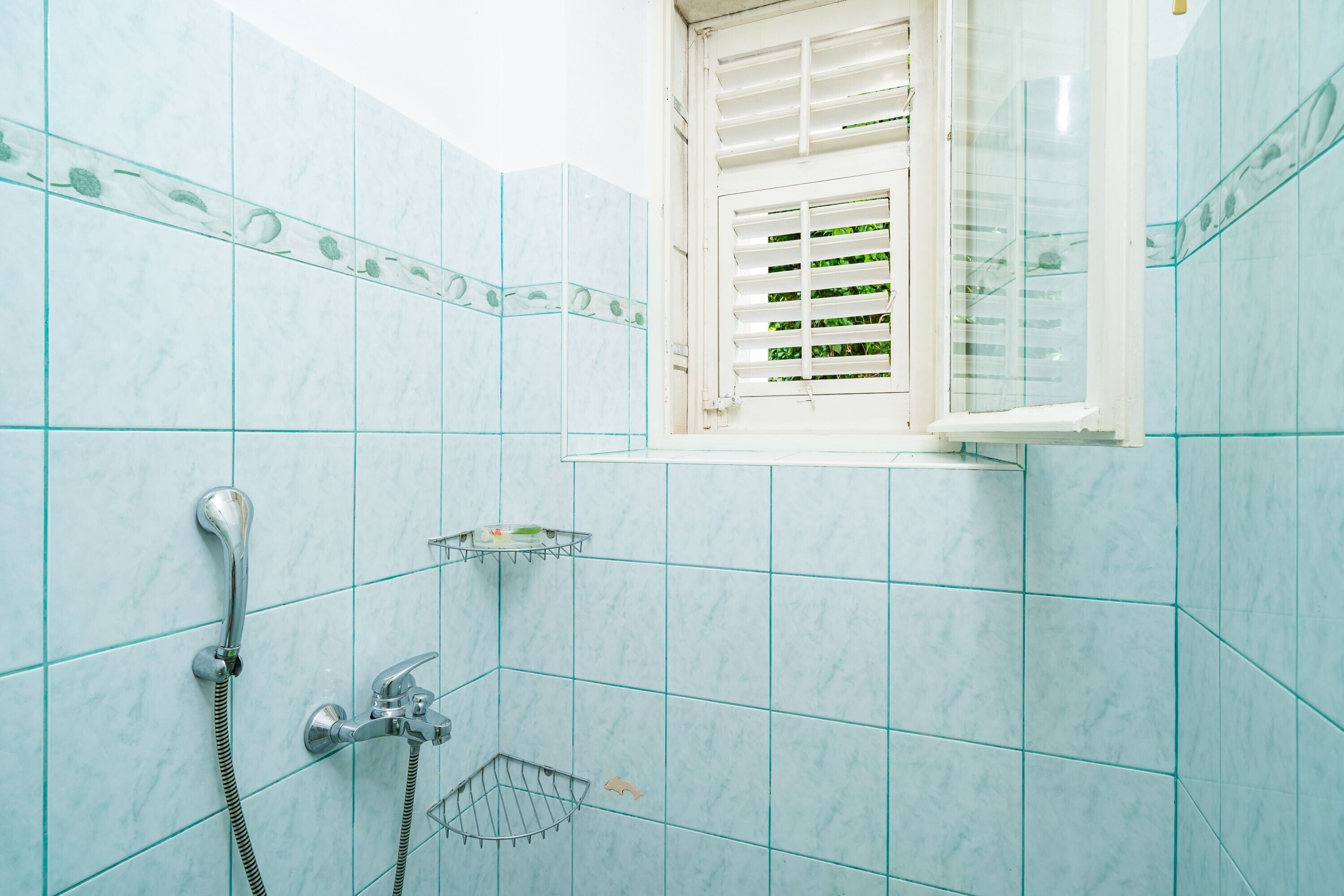 Une salle de bain avec une douche, une fenêtre avec des stores et une petite étagère.