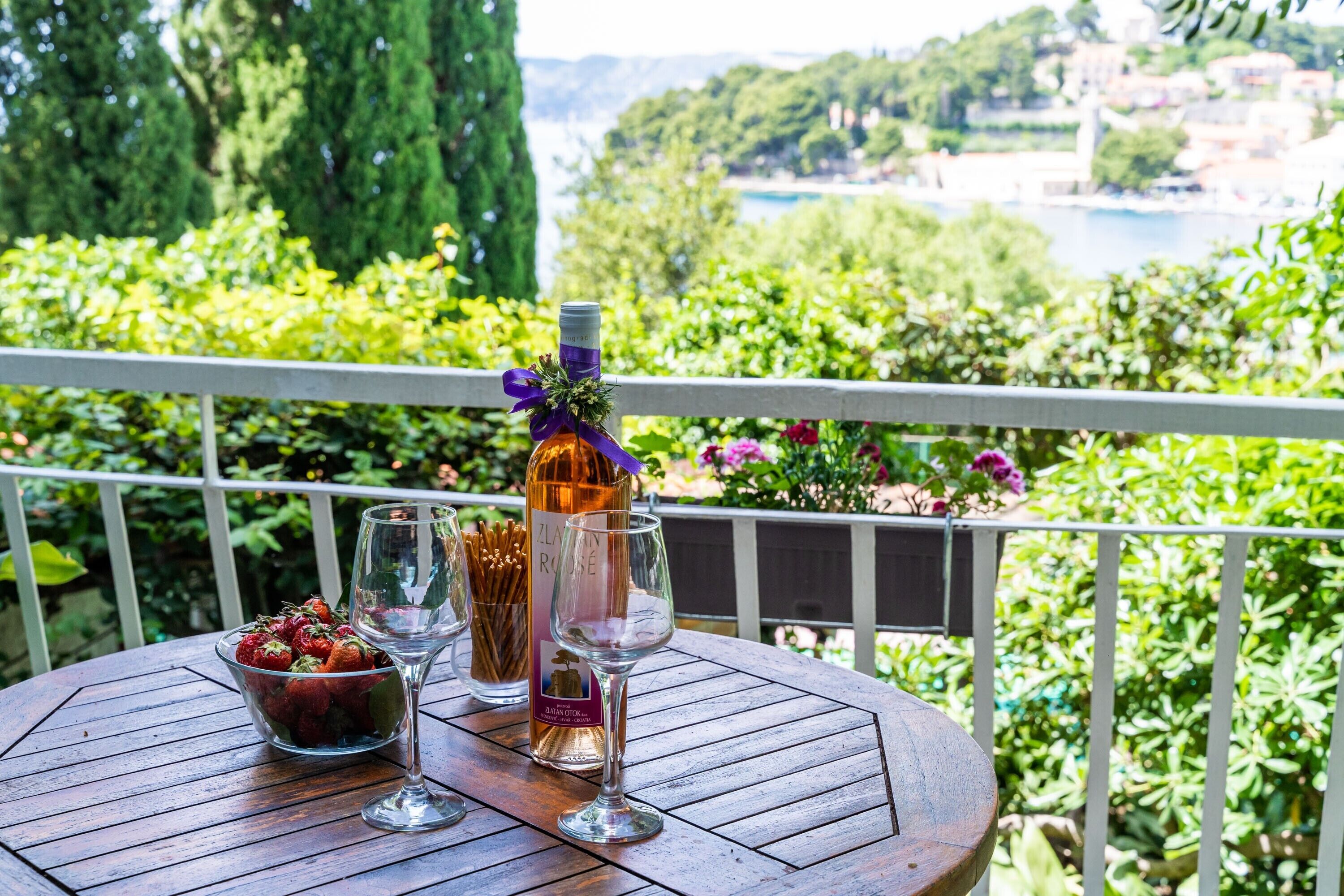 Une table ronde en bois, sur laquelle sont posés une bouteille de vin, deux verres et un bol de fraises, se trouve sur un balcon offrant une vue panoramique.