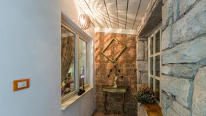 A narrow hallway with a stone wall, a window with blinds, a small table with a vase, and a wall-mounted decorative item.