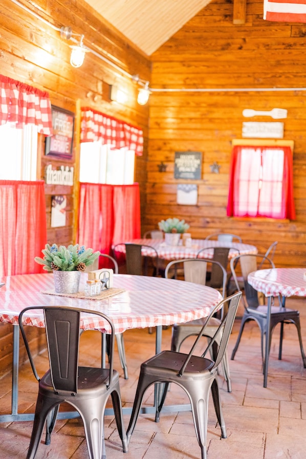 A rustic wooden interior with round tables and metal chairs, red curtains, and a small potted plant on one table.