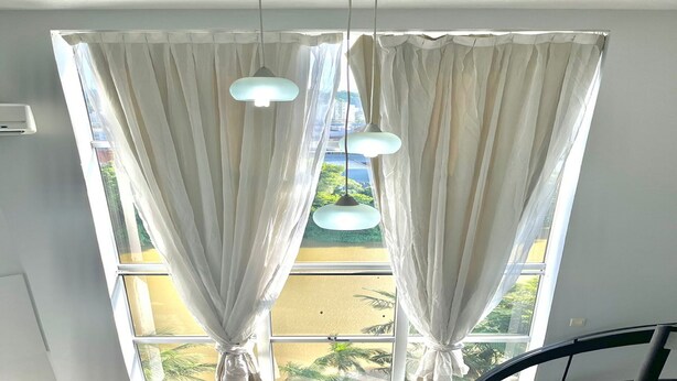 Una habitación con un ventanal grande, cortinas blancas y dos lámparas colgantes.