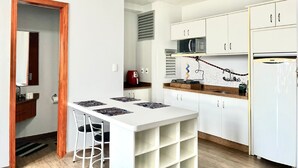 Cocina compacta con armarios blancos, microondas y refrigerador. Cuenta con un pequeño comedor con mesa y sillas.