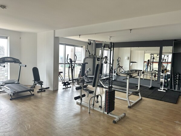Un gimnasio espacioso con diversos equipos de ejercicio, ventanas amplias y un espejo que refleja el espacio.