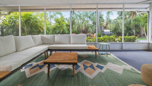 Uma sala de estar moderna com um sofá seccional branco, uma mesa de centro de madeira e uma pequena mesa redonda. O espaço possui grandes janelas com vista para vegetação exuberante e uma churrasqueira do lado de fora.