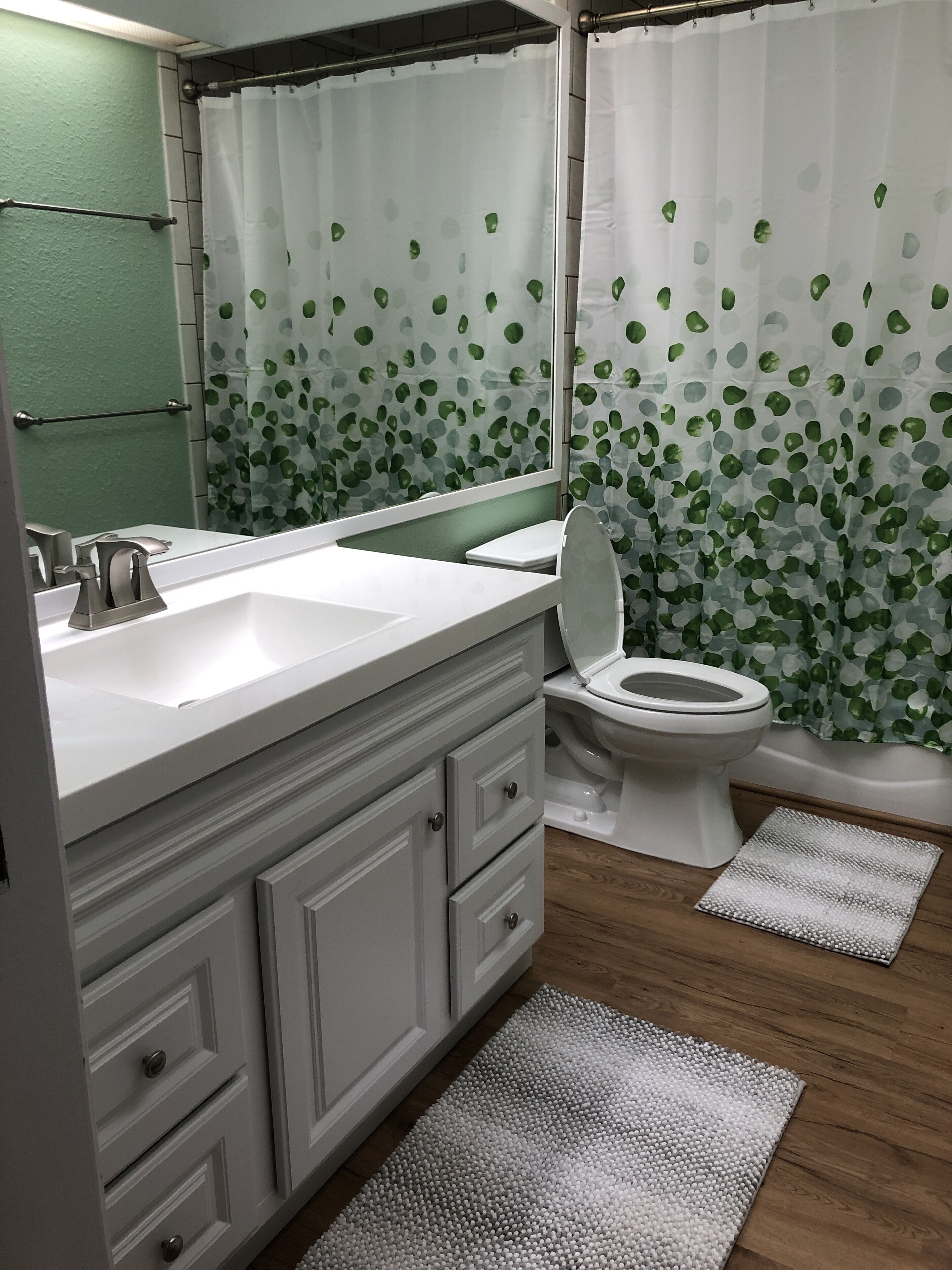A bathroom with a white sink, mirror, and toilet. There is a green shower curtain with leaf patterns.