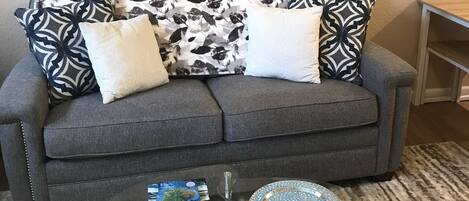 Una sala de estar con un sofá gris, cojines estampados, una mesa de centro de vidrio con un libro y un vaso, y dos cuadros en la pared.