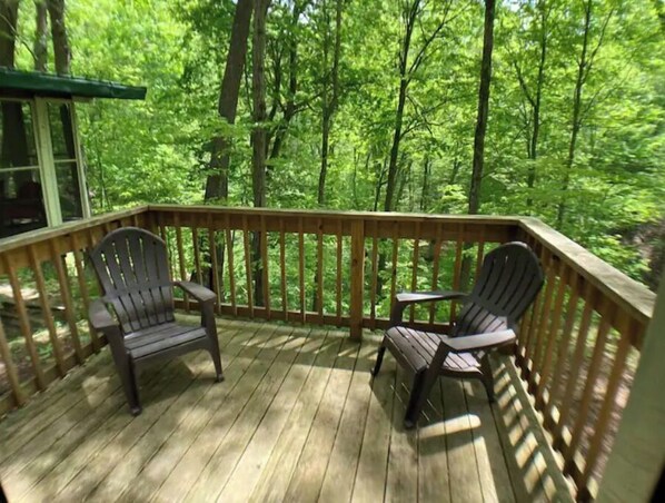 A wooden deck with two Adirondack chairs and a small table, surrounded by a wooden railing, overlooking a forested area. - Treetops Cabin in the Woods! (Bloomingdale)