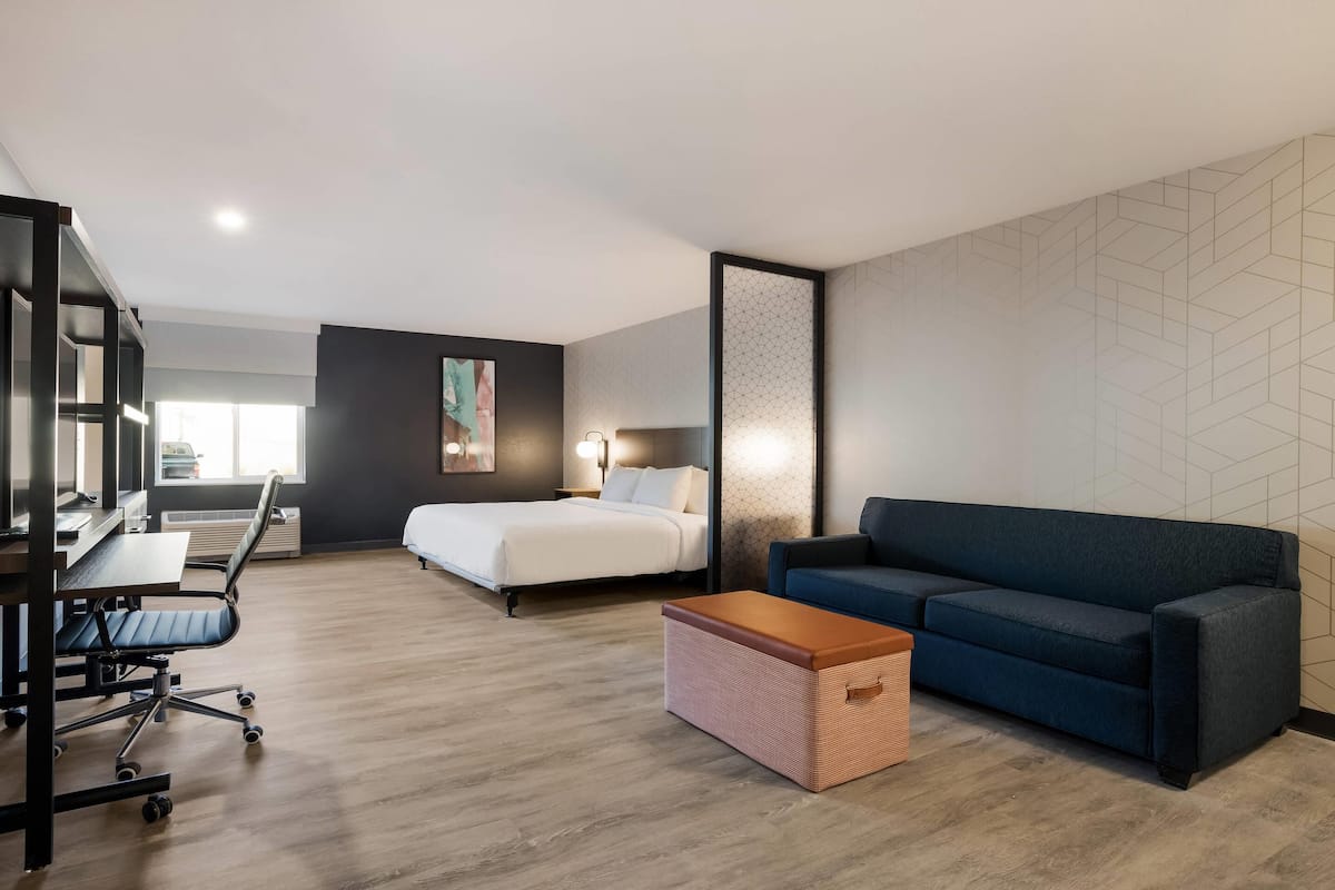 A modern hotel room with a bed, a blue sofa, a desk with a chair, and a small wooden chest.