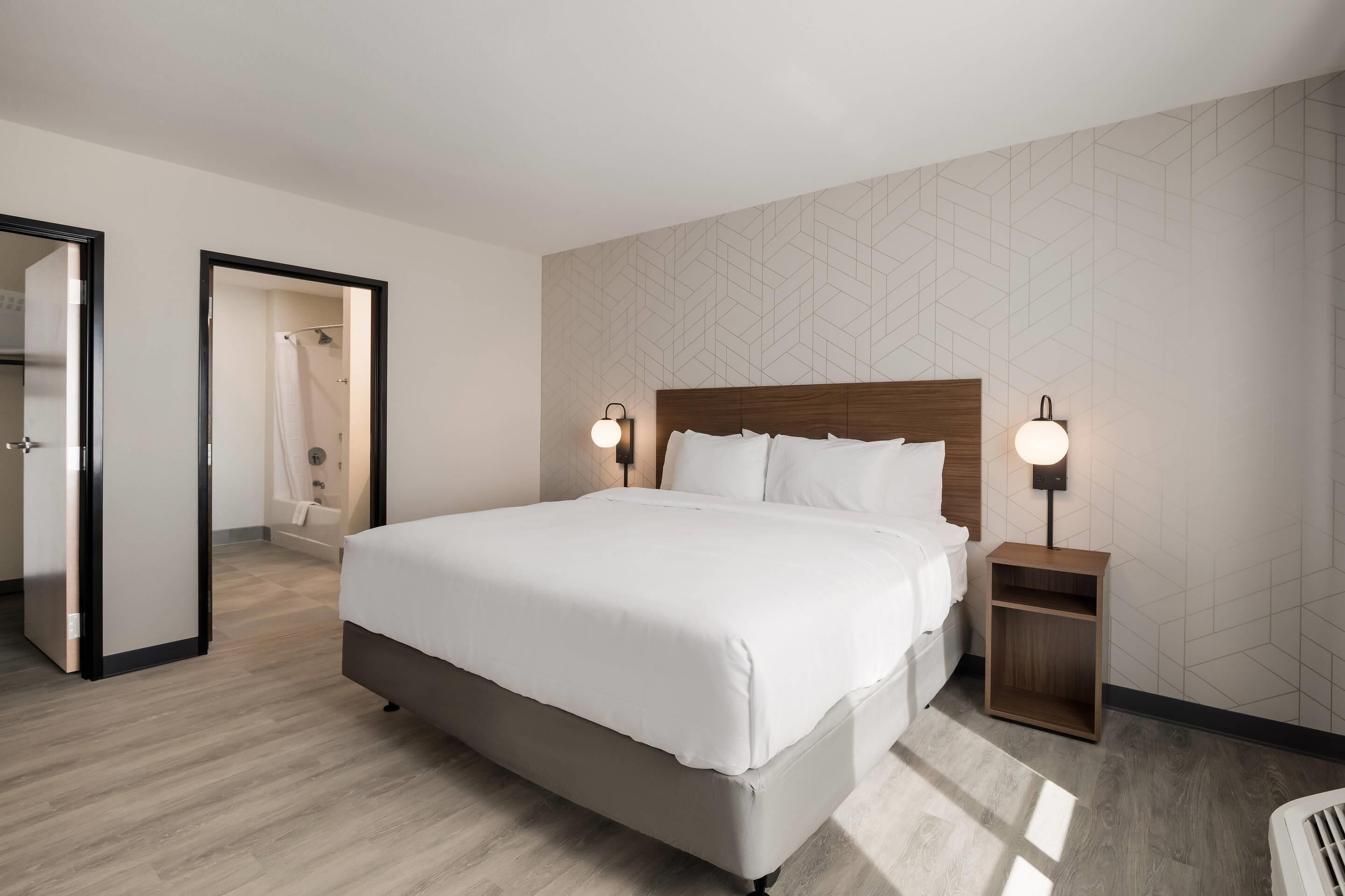 Une chambre d’hôtel moderne avec un grand lit, deux tables de chevet avec des lampes, une tête de lit en bois et un mur orné d’un motif géométrique.