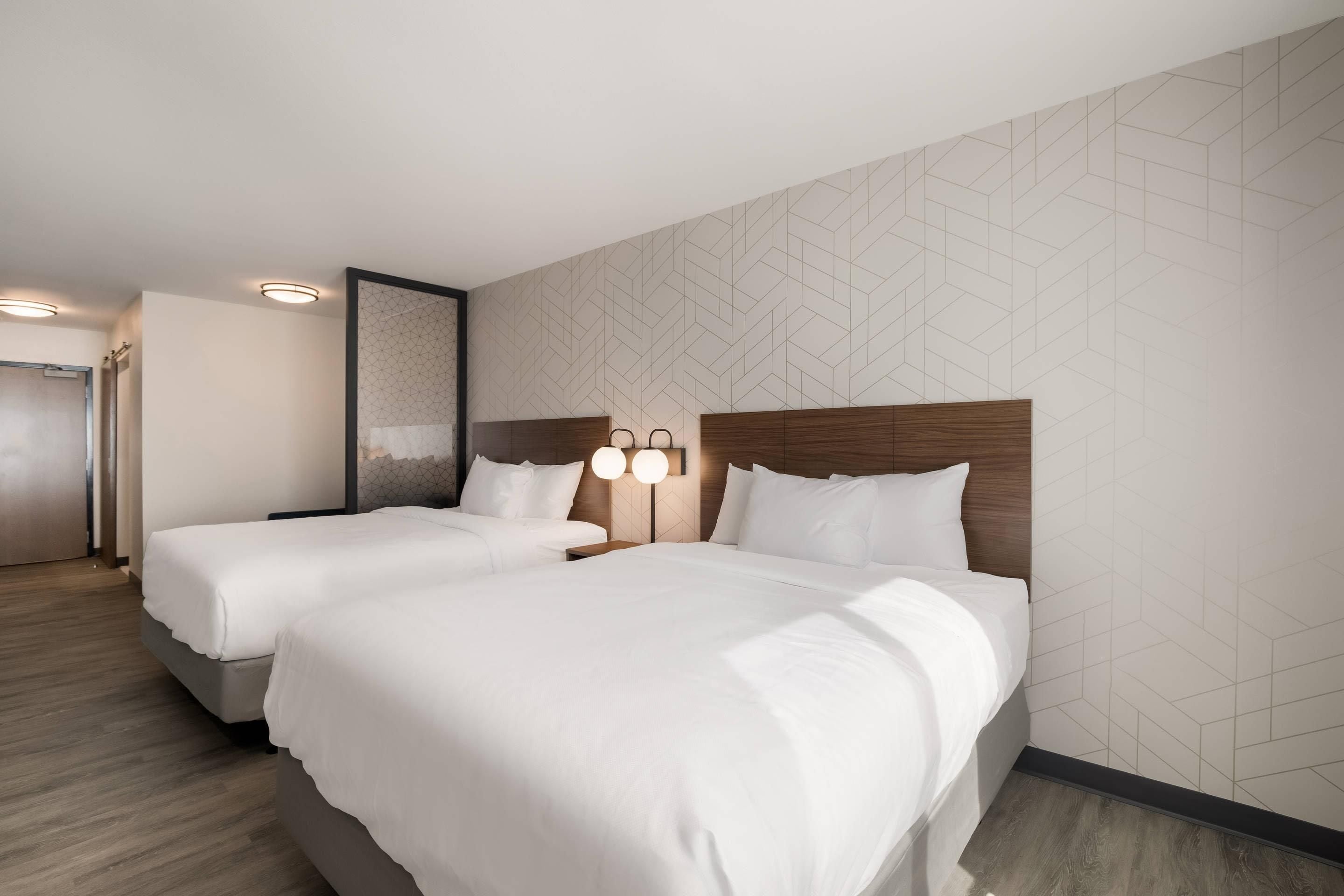 Quarto de hotel com duas camas, cabeceira de madeira, luminária fixada na parede e parede com padrão geométrico.