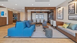 A modern living area with a blue sofa, a grey sofa, and a round table. There are bookshelves and a dining area in the background. - Everhome Suites Corona (Corona)