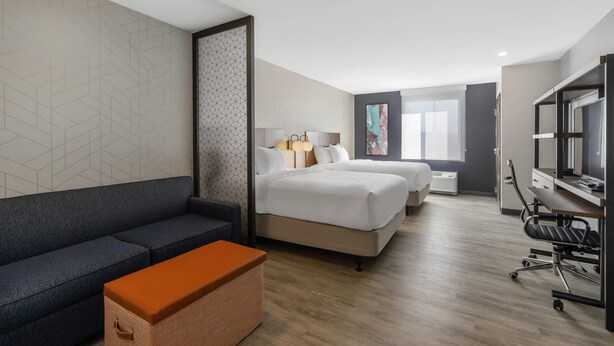 A hotel room with a sofa, two beds, a desk, and a TV. - Everhome Suites Corona (Corona)