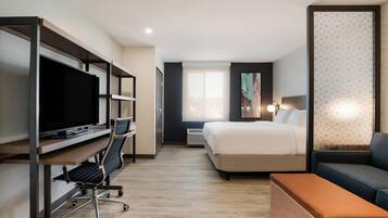 A modern hotel room with a bed, a desk with a computer, a TV, and a seating area with a sofa and ottoman.