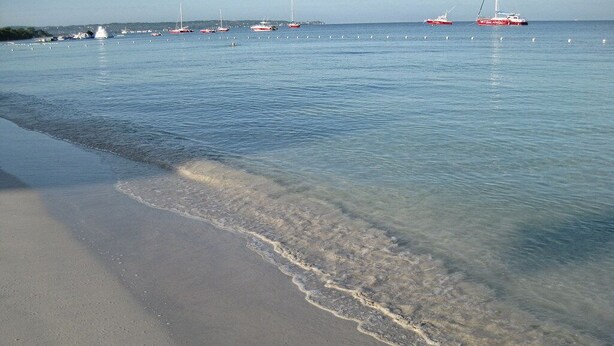 Een strand met helder water en boten in de verte.